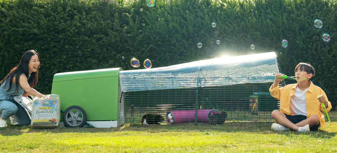 Een kind dat bellen blaast in de tuin, terwijl zijn moeder hooi ververst in het hok Eglu Go .