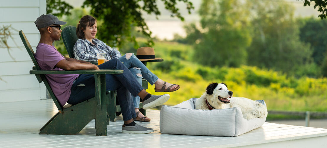 Hond rustend op hondenbed van ribfluweel met kiezelsteentjes op veranda, met baasjes achter hen op houten stoelen.